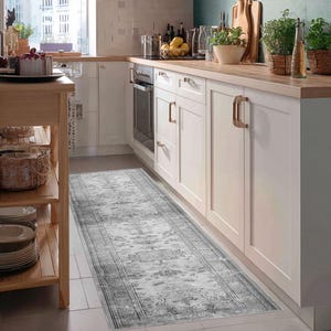 Könnte beinhalten: Ein rechteckiger Läuferteppich mit Used-Look in einer Küche. Der Teppich hat ein verblasstes grau-weißes Muster mit einem Bordürendesign. Die Küche hat weiße Schränke, eine Holz-Arbeitsplatte und ein Fenster.
