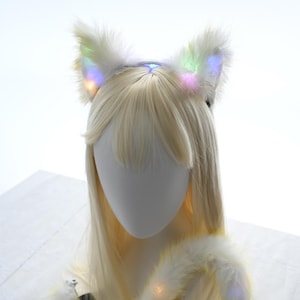 May include: White cat ear headband with fluffy, light-up ears. The ears have a rainbow color effect. The headband is on a mannequin head with long, blonde hair. The mannequin is wearing a white furry accessory.