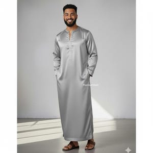 May include: A long, silver-gray thobe with long sleeves and a mandarin collar. The traditional garment has a relaxed fit and pockets. The text "PremiumVastr" is visible on the garment.