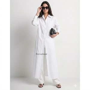 Può includere: Un lungo abito camicia bianco con maniche lunghe e spacco frontale, abbinato a pantaloni bianchi e sandali neri. La modella indossa occhiali da sole e tiene in mano una pochette nera. Il testo "PremiumVastr" è visibile.