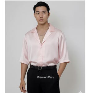 May include: A light pink satin shirt with short sleeves and a classic collar. The shirt is buttoned down and paired with black pants and a black belt. The text "PremiumVastr" is visible on the pants.