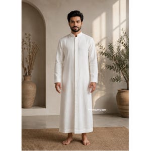 May include: A long-sleeved, floor-length white thobe featuring a mandarin collar and a concealed front button placket. The garment has a straight-cut silhouette and plain cuffs. The text "PremiumVastr" is visible near the bottom right hem.