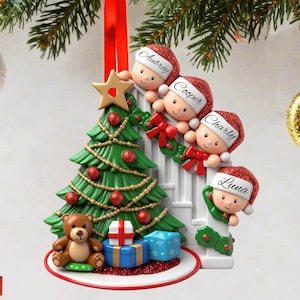 Könnte beinhalten: Weihnachtsschmuck mit einem grünen Weihnachtsbaum, roten Kugeln und einem Teddybären. Vier Figuren mit Weihnachtsmützen tragen Namen. Ein goldener Stern und ein rotes Band schmücken die Oberseite. Geschenke befinden sich am Fuß des Baumes.