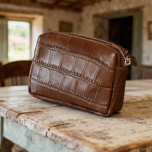May include: A brown, rectangular cosmetic bag with a crocodile skin texture. The bag has a gold zipper closure and a small pull tab. The bag is sitting on a distressed wooden table.