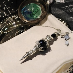 K&ouml;nnte beinhalten: Ein silberfarbenes Pendel mit einem schwarzen Edelstein und einem Sternanh&auml;nger, das auf einem wei&szlig;en Tuch liegt. Das Pendel hat eine Spitze und Zierdetails. Eine silberne Schale und ein Glas mit gr&uuml;nem und blauem Design sind im Hintergrund.