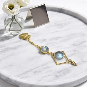 Könnte beinhalten: Eine goldfarbene Halskette mit zwei blau getönten Edelsteinen und einem quadratischen Anhänger. Die Kette liegt auf einem weißen Marmortablett mit einer kleinen Glasvase mit weißen Blumen.