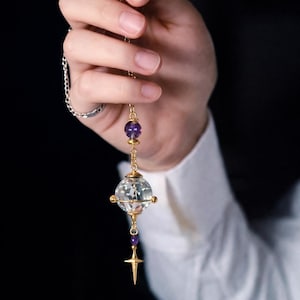 May include: A gold pendant necklace with a clear crystal orb, purple amethyst beads, and a gold cross charm. The necklace hangs from a fine chain. The design is elegant and detailed, with a focus on the gemstones and metalwork. The necklace is a piece of jewellery.