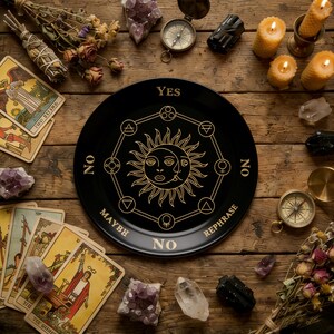 Può includere: Un piatto di divinazione nero con simboli e testo dorati, tra cui "SÌ", "NO" e "FORSE". Il piatto presenta un disegno di sole e luna. Tarocchi, cristalli, candele e una bussola circondano il piatto su una superficie di legno.