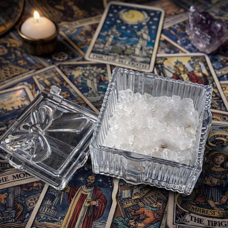 K&ouml;nnte beinhalten: Ein klarer, quadratischer Glasbeh&auml;lter gef&uuml;llt mit wei&szlig;en Kristallen, neben seinem Deckel mit einem Schleifendesign. Der Beh&auml;lter steht auf einer Oberfl&auml;che, die mit Tarotkarten bedeckt ist, mit einer brennenden Kerze und einem lila Kristall im Hintergrund.