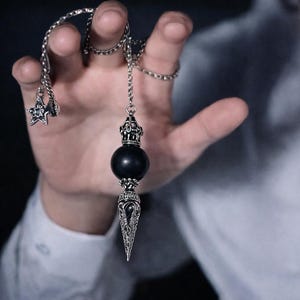 Op de afbeelding: Een zilverkleurige pendel met een zwarte bol en een decoratief, puntig uiteinde. De pendel hangt aan een ketting met een sterbedel. Het ontwerp heeft ingewikkelde details en wordt in een hand gehouden tegen een donkere achtergrond.