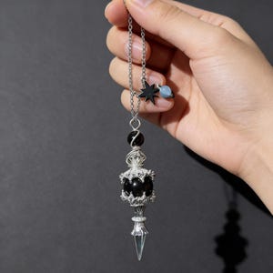 May include: A silver-toned pendulum with a black sphere, star, and blue bead accents. The pendulum has a clear, pointed crystal at the bottom and is suspended from a chain. The design incorporates celestial elements.