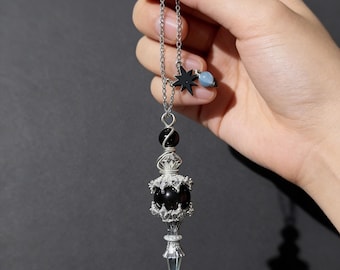 May include: A silver-toned pendulum with a black sphere, star, and blue bead accents. The pendulum has a clear, pointed crystal at the bottom and is suspended from a chain. The design incorporates celestial elements.