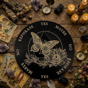 May include: A black divination board featuring a gold butterfly and floral design, with the words "Yes," "Maybe," "No," and "Rephrase." The image also shows tarot cards, crystals, candles, and compasses, arranged on a wooden surface.