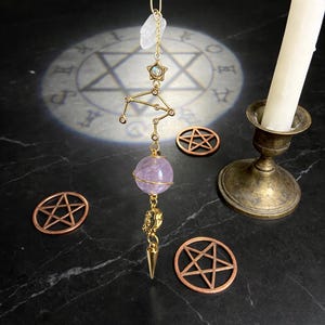 Puede incluir: Un colgante dorado del signo zodiacal Libra con una esfera de amatista púrpura y un cristal de cuarzo transparente. El colgante cuelga con pentagramas de cobre y un candelabro de latón con una vela blanca encendida. El fondo es negro.