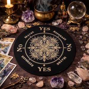Könnte beinhalten: Ein schwarzes Wahrsagebrett mit goldfarbenen Buchstaben und einem zentralen floralen Design. Das Brett enthält die Wörter "YES", "NO", "MAYBE" und "REPHRASE". Tarotkarten und Kristalle umgeben das Brett.