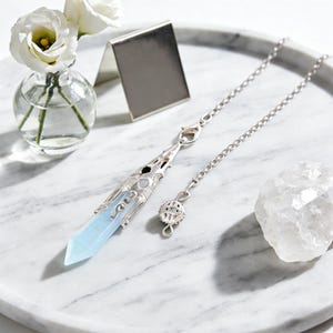 May include: A silver pendulum with a light blue crystal point and a decorative chain. The pendulum is displayed on a white marble tray, alongside a small vase with white flowers and a clear quartz crystal.