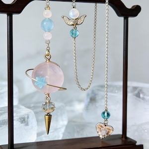 May include: Earrings with celestial-themed charms. One earring features a pink planet with a blue star and gold accents. Another has a gold angel wing charm and a blue bead. A third earring has a heart charm.