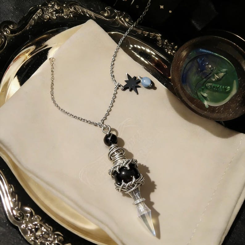 K&ouml;nnte beinhalten: Eine silberfarbene Anh&auml;ngerkette mit einer schwarzen, drahtumwickelten Perle, einer Spitze aus klarem Kristall und einem Stern- und blauen Perlenanh&auml;nger. Die Kette wird auf einem wei&szlig;en Tuch pr&auml;sentiert.