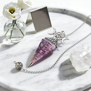 May include: A purple crystal pendulum with a silver chain and decorative top featuring a pentagram and crescent moons. The pendulum is on a white marble tray, next to a small glass vase with white flowers and a clear quartz crystal.
