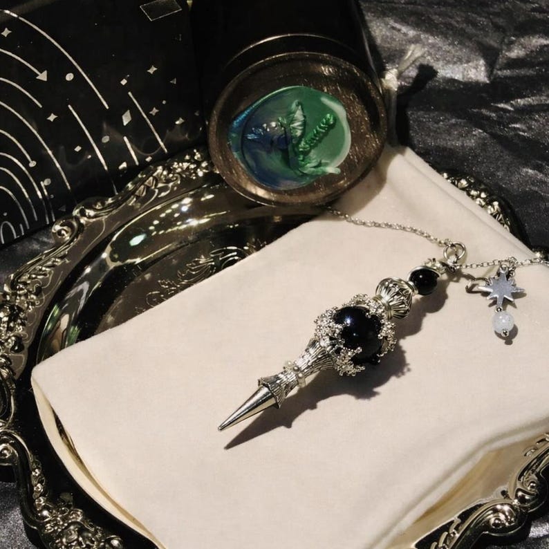 K&ouml;nnte beinhalten: Ein silberfarbenes Pendel mit einem schwarzen Edelstein und einem Sternanh&auml;nger, das auf einem wei&szlig;en Tuch liegt. Das Pendel hat eine Spitze und Zierdetails. Eine silberne Schale und ein Glas mit gr&uuml;nem und blauem Design sind im Hintergrund.