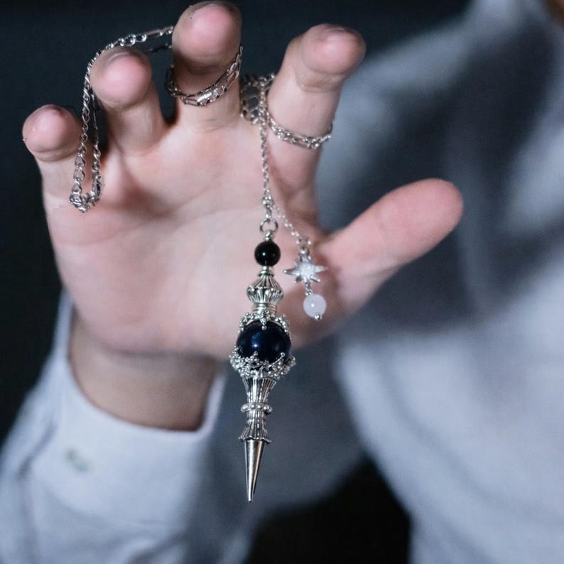 K&ouml;nnte beinhalten: Eine silberfarbene Pendelkette mit einem dunkelblauen Orb, einer schwarzen Perle und einem sternf&ouml;rmigen Anh&auml;nger. Das Pendel hat ein kronenartiges Design und eine spitze Spitze. Die Kette wird von einer Hand gehalten.