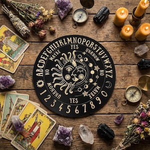May include: A black planchette featuring a sun and moon design, alongside tarot cards, crystals, candles, and compasses. The planchette is inscribed with "YES", "NO", "MAYBE", and "UNKNOWN", plus the alphabet and numbers. The items are arranged on a wooden surface.