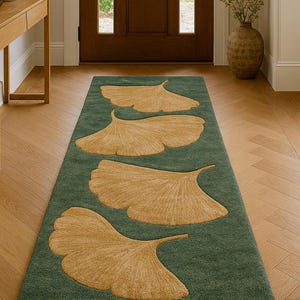 May include: A green rectangular rug featuring four large, tan-coloured ginkgo leaf designs. The rug is in a hallway with a wooden door and light-coloured walls. The floor is made of light-coloured wood.