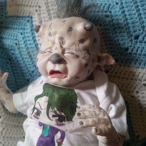May include: A detailed, realistic doll with a unique, creature-like design. The doll has a light skin tone with dark spots and a distressed expression. It wears a white t-shirt featuring a cartoon graphic of a character. The doll is lying on a blue and white crocheted blanket.