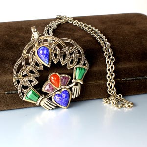May include: Silver-toned Claddagh pendant necklace with a Celtic knot design. The pendant features colorful gemstone accents in blue, red, green, and purple. The necklace has a delicate chain.
