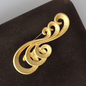 May include: A gold-toned brooch featuring a series of interconnected, textured swirls and curved lines. The metal surface displays a brushed finish with visible striations throughout the design, creating a continuous, flowing, and abstract shape.