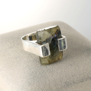 May include: A silver ring featuring a rectangular, faceted labradorite gemstone with dark green and grey tones. The stone is held in place by a wide, polished silver band that wraps around the sides, creating a bold, geometric look for the finger.