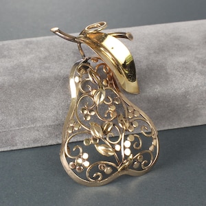 May include: A gold-toned pear-shaped brooch with intricate openwork filigree design. The brooch features a solid leaf and stem, and the pear shape is filled with floral and vine details. The brooch is likely made of metal.