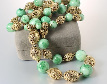 May include: A long green and gold beaded necklace with a filigree design. The necklace is on a grey velvet box.