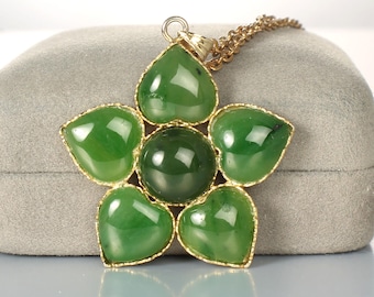May include: A gold-toned necklace featuring a flower-shaped pendant. The pendant has a central, round, dark green stone surrounded by five heart-shaped, lighter green stones. The stones are set in a gold-colored metal frame.