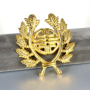 May include: A gold-colored brooch with a central circular design and a surrounding laurel wreath. The brooch has a detailed, textured surface and is set against a gray background. The design is symmetrical and ornate.