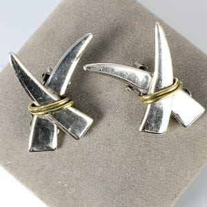 May include: A pair of silver earrings with a gold accent. The earrings are shaped like two crossed horns.