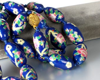 May include: A close-up of a blue beaded necklace with floral designs. The oval-shaped beads are a deep blue with hand-painted pink, white, and green floral patterns. The necklace is strung on blue cord and has a gold-colored clasp.