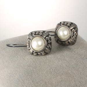 May include: A pair of silver-toned earrings with a square design. Each earring features a central, round, white pearl surrounded by ornate filigree detailing. The earrings are displayed on a gray surface, showcasing their intricate design and elegant appeal.