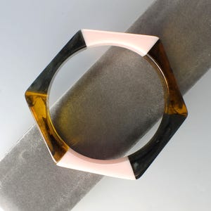 May include: A hexagonal bangle bracelet featuring alternating segments of solid pale pink resin and translucent amber-brown tortoiseshell-patterned resin. The geometric design has a smooth, polished finish with a circular inner opening for the wrist.