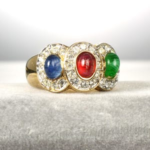 May include: A gold ring featuring three oval gemstones: a blue sapphire, a red ruby, and a green emerald. Each gemstone is surrounded by a halo of small, clear crystals. The ring has a classic, elegant design.