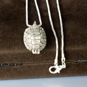 May include: A silver-toned turtle pendant necklace featuring a detailed shell design on a thin snake chain. The pendant is shaped like a turtle with a textured carapace and a small head, attached to a metallic chain with a lobster claw clasp.