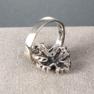 Peut inclure: Une bague en argent avec un design orn&eacute; et d&eacute;taill&eacute;. La bague pr&eacute;sente une pierre centrale de couleur fonc&eacute;e entour&eacute;e d'un sertissage d&eacute;coratif. Le bandeau est lisse et poli, contrastant avec le design textur&eacute; de la face de la bague.