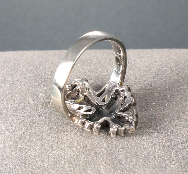 Peut inclure: Une bague en argent avec un design orn&eacute; et d&eacute;taill&eacute;. La bague pr&eacute;sente une pierre centrale de couleur fonc&eacute;e entour&eacute;e d'un sertissage d&eacute;coratif. Le bandeau est lisse et poli, contrastant avec le design textur&eacute; de la face de la bague.