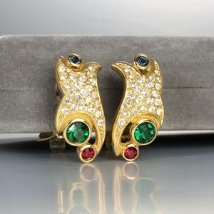 May include: Gold tone clip-on earrings with a swirl design. The earrings are encrusted with clear rhinestones and feature green and red stones.