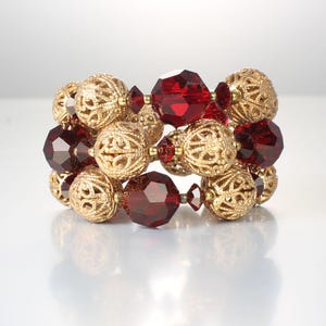 May include: A gold-colored beaded bracelet with filigree spheres and faceted red beads. The bracelet is made of multiple strands of beads, creating a layered effect. The beads are arranged in a repeating pattern of gold and red.