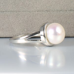 May include: A silver ring featuring a large, lustrous white pearl. The pearl has a subtle pink iridescence. The ring's band has a split design, with the pearl held in a smooth, rounded setting. The ring is displayed on a reflective surface.