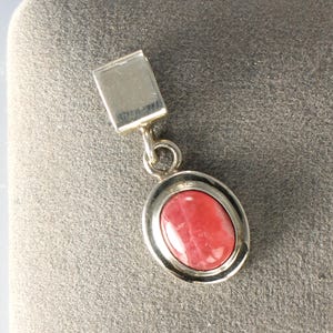 May include: A silver pendant featuring an oval, red gemstone. The gemstone is set in a silver bezel and suspended from a rectangular silver bail. The pendant is shown against a grey backdrop.