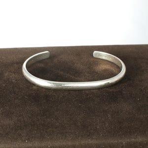 May include: A silver-colored metal cuff bracelet with a smooth, polished finish and a rounded profile. The bracelet features an open-ended design, allowing for adjustment, and includes small, engraved markings on the inner surface near the ends.