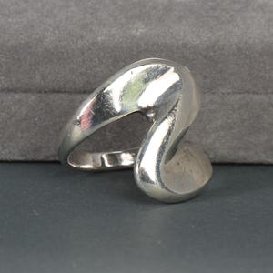 May include: A polished silver-toned ring featuring a continuous, fluid, and asymmetrical wave-like design. The band curves into a prominent, sculptural loop that creates an open, abstract shape with smooth, reflective surfaces throughout the piece.