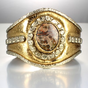 May include: A gold and silver cuff bracelet with a large, round, brown and white agate gemstone set in the center. The bracelet has a textured surface and a beaded border.